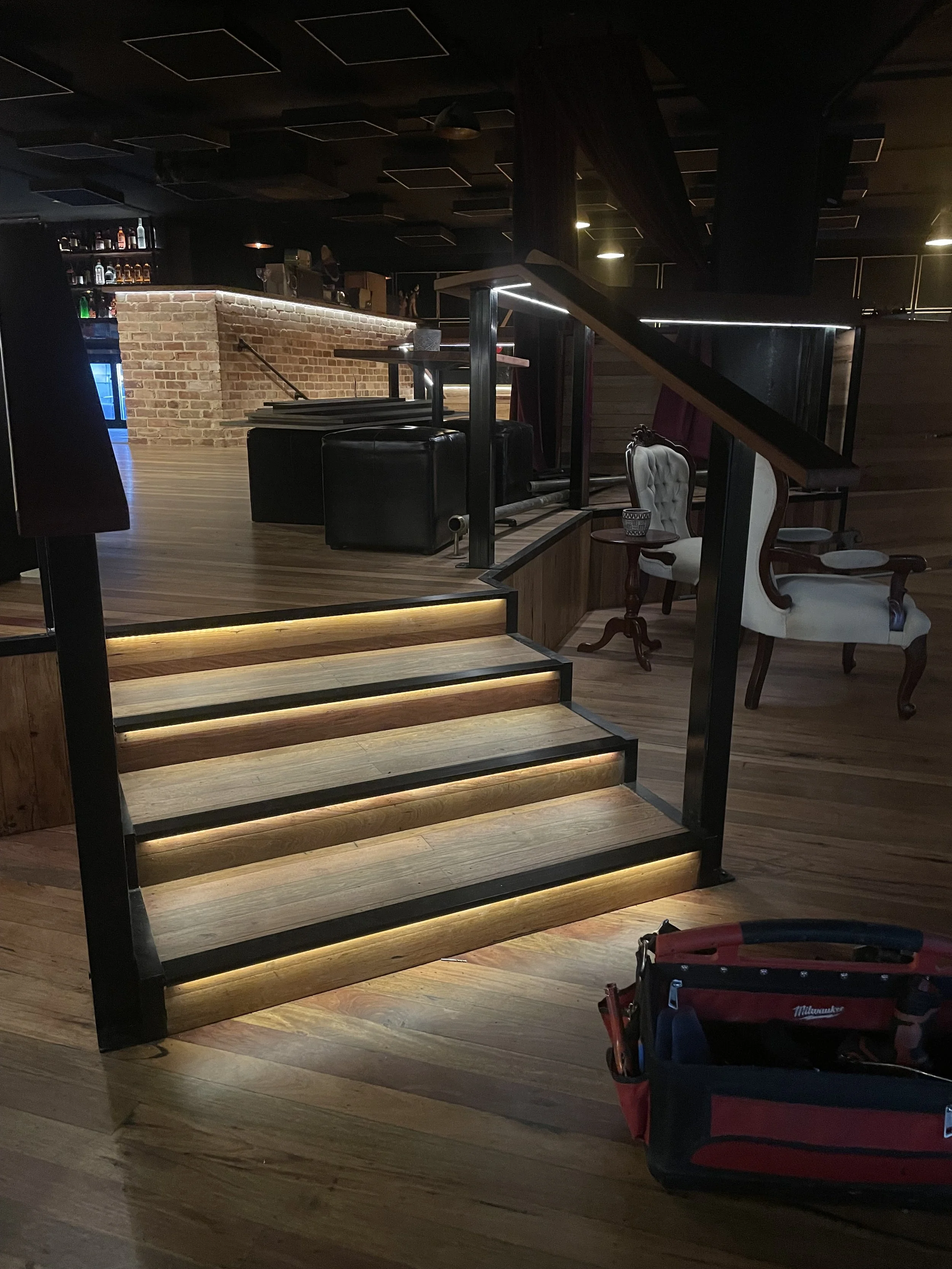 Dimly lit interior of a bar or lounge with a brick wall bar counter, wooden floors, and staircase with illuminated steps. There are tables, chairs, and some bottles on shelves in the background. A red toolbox is in the lower right corner.