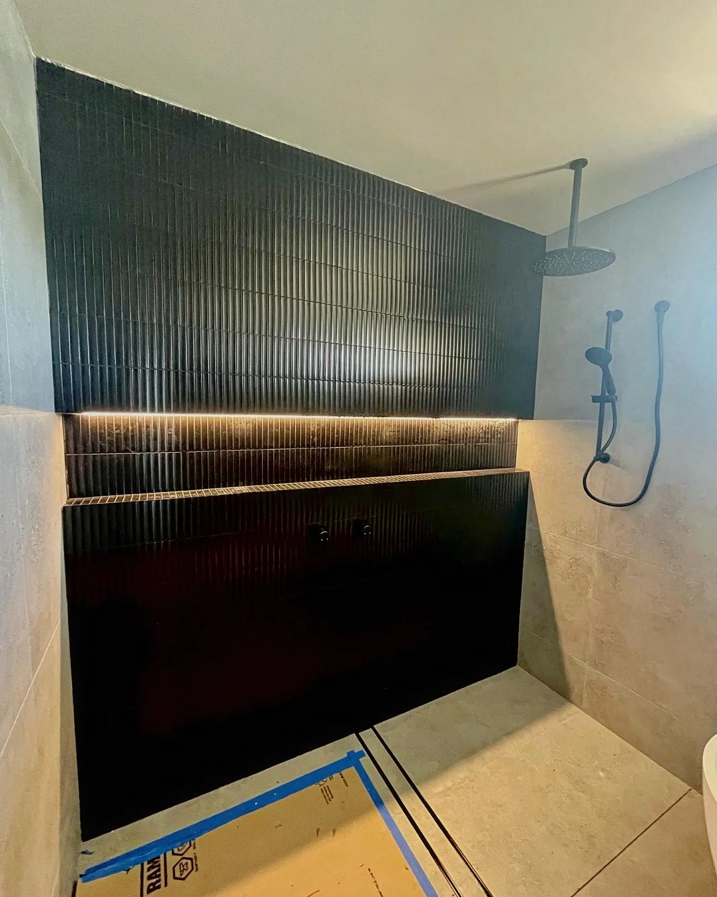A modern walk-in shower with a black tiled wall, a rainfall showerhead, and a handheld shower wand, with beige tiles on the adjacent wall and a strip of built-in lighting.