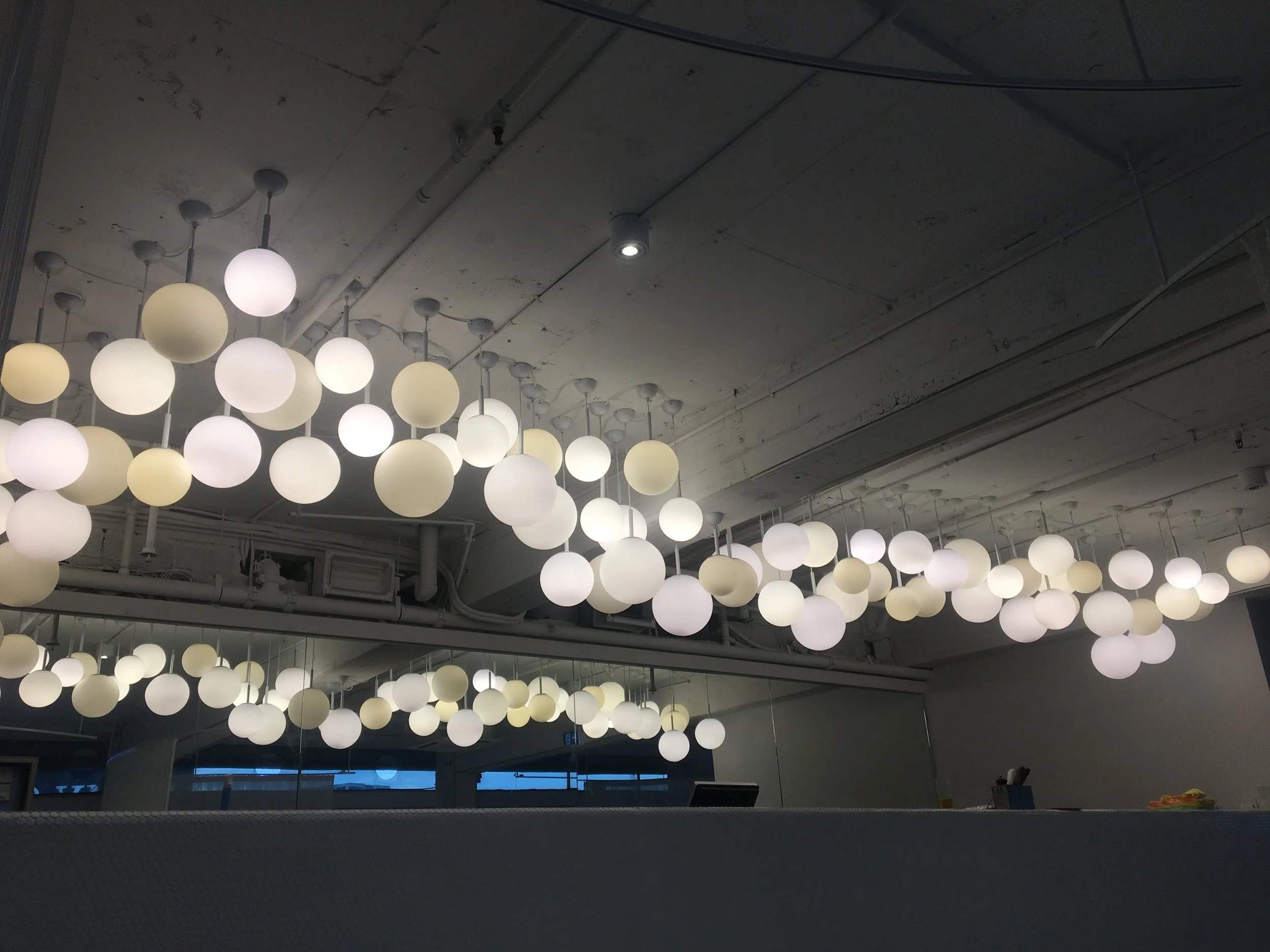 Ceiling with numerous spherical pendant lights hanging in rows, some illuminated softly, in an indoor space with an industrial-style ceiling featuring pipes and exposed wiring.