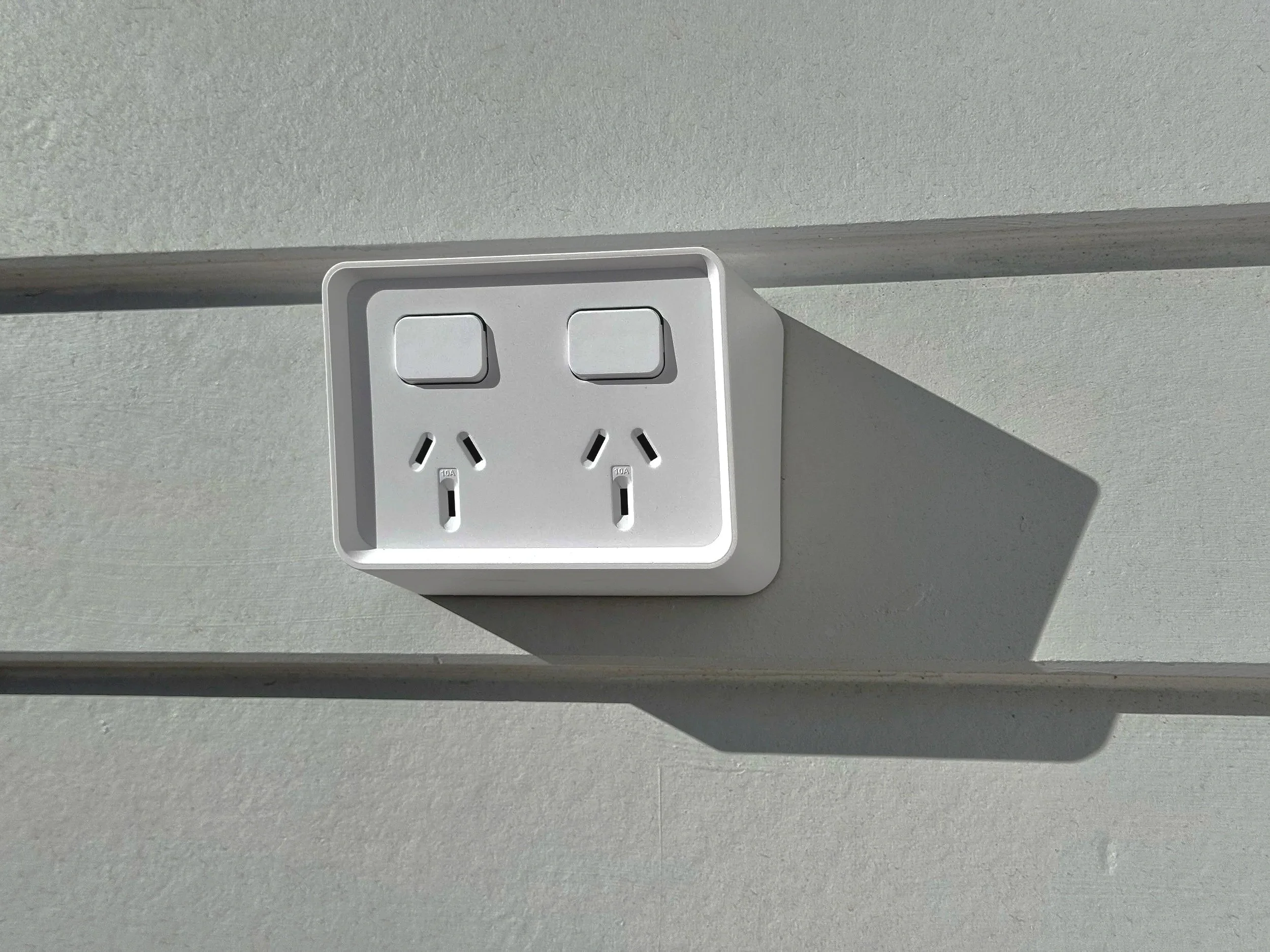 A white clipsal iconic electrical weatherproof outlet on a light gray wall, with two sockets casting a shadow to the right.
