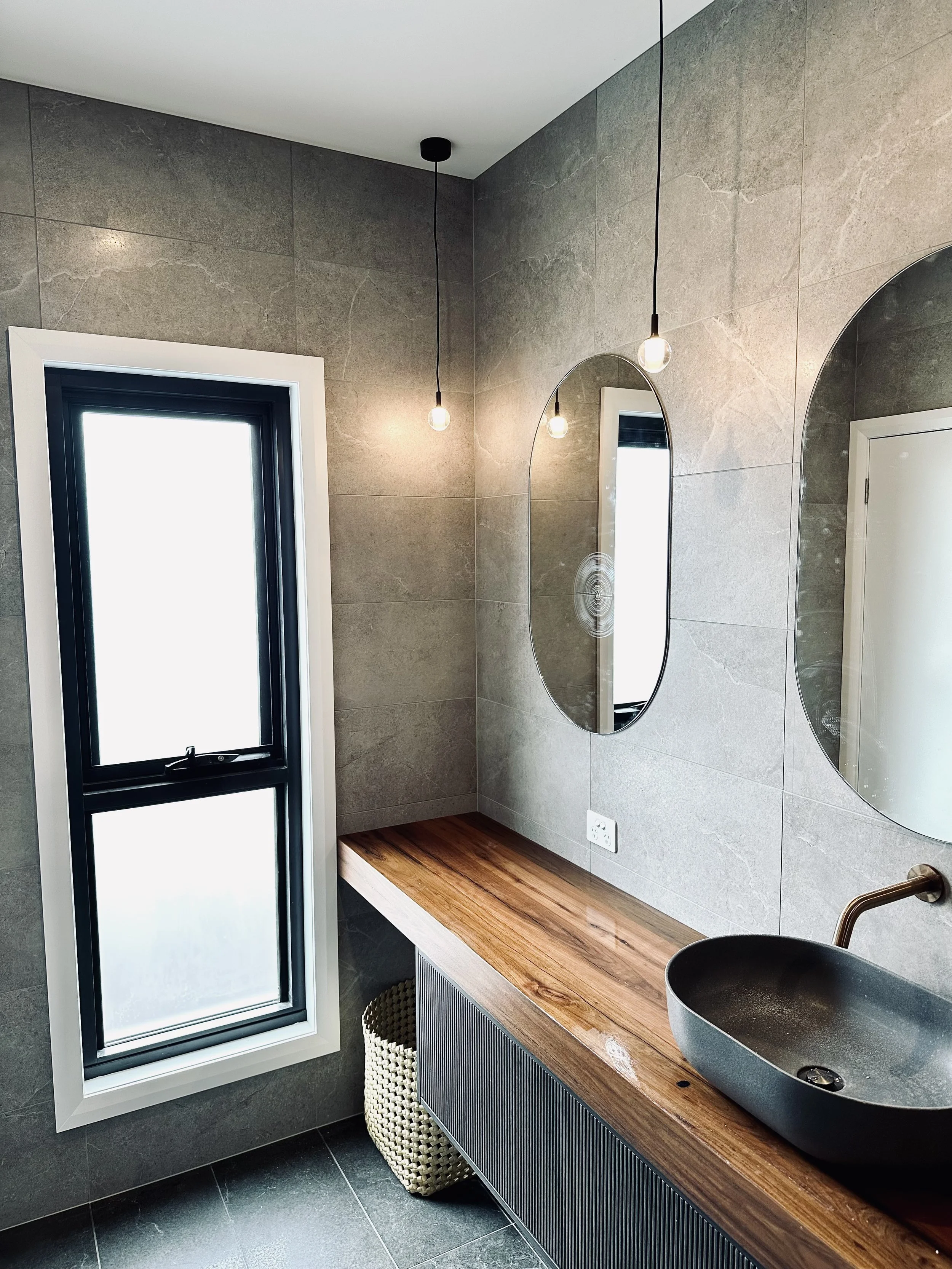 Modern bathroom with a window, oval mirror, hanging pendant lights, wooden countertop, and a black vessel sink.