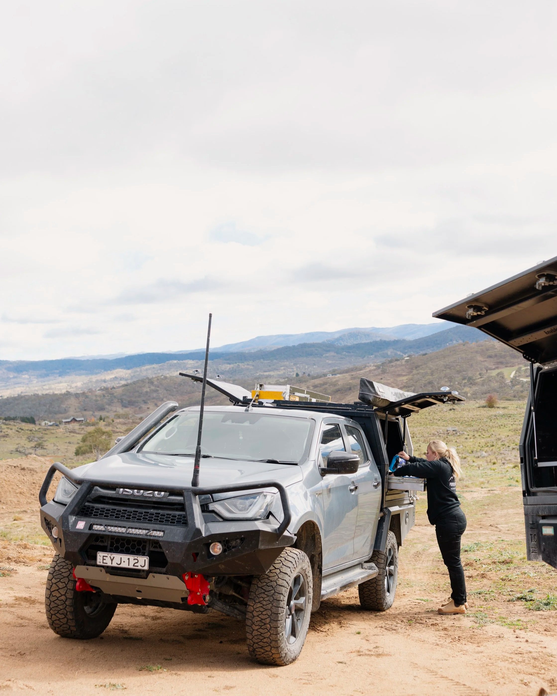 A woman in black pants and boots is working on a silver off-road vehicle with a bull bar and large tires, parked on a dirt field under a cloudy sky with mountains in the background.