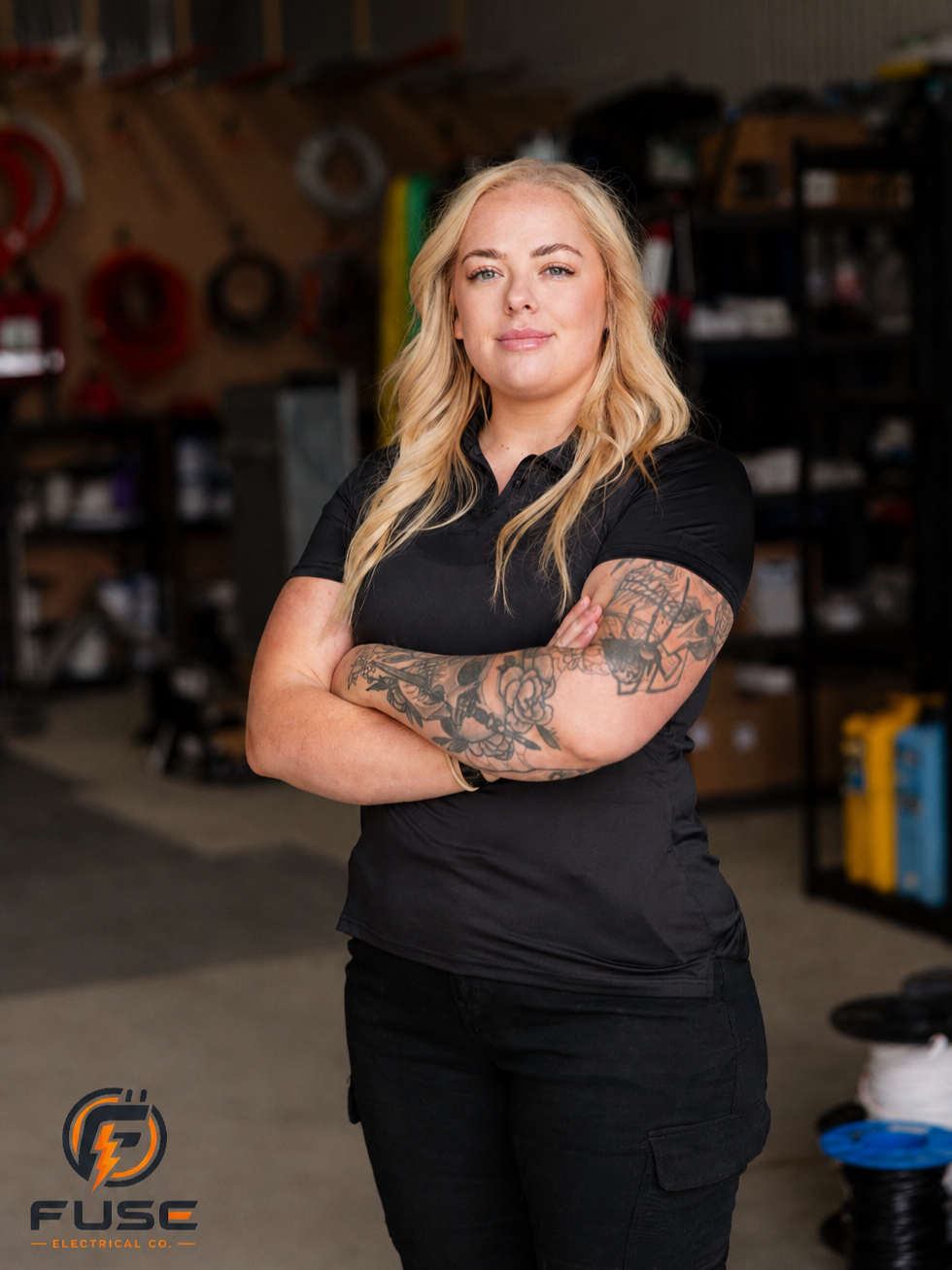 A confident woman with blonde hair, tattoos on her arms, and wearing a black polo shirt stands with arms crossed in a workshop or hardware store, with various tools and hardware supplies in the background.