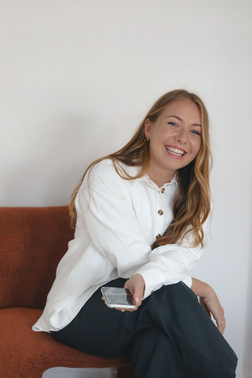 Hayley McAllister, Founder of Groundset HR, the HRIS Implementation Specialists, with long red hair smiling, sitting on a brown chair, holding a smartphone, wearing a white button-up shirt and black pants, against a plain white wall.