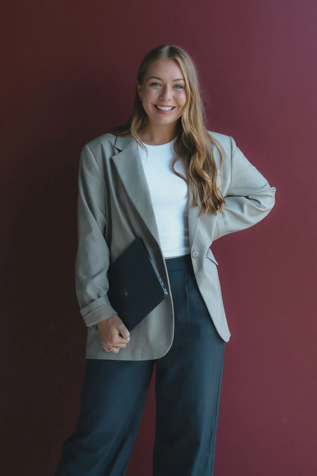 A woman with long, wavy blonde hair smiling, wearing a light grey blazer, white top, and dark trousers, holding a laptop in her left hand, standing against a maroon background.