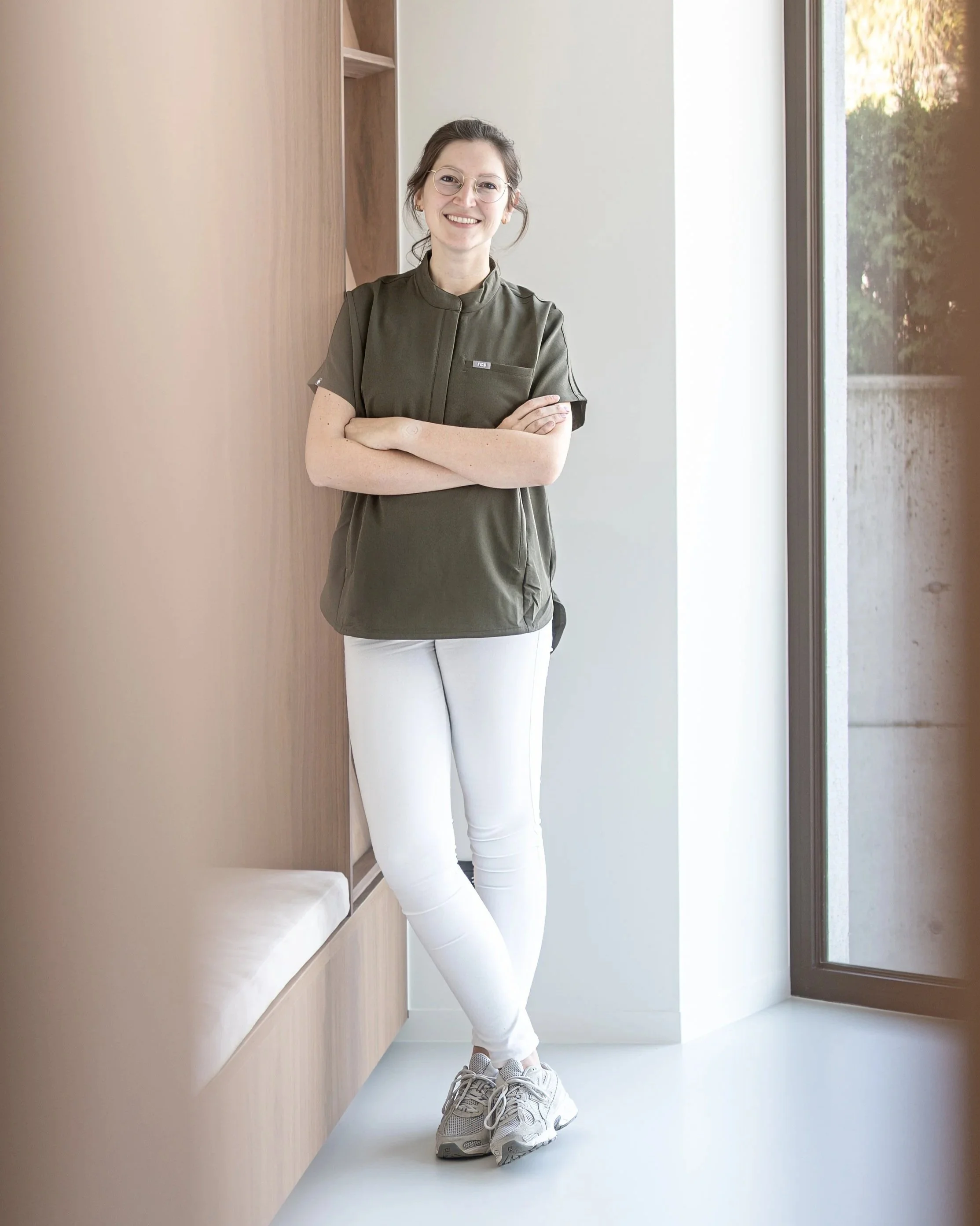 Vrouw met bril, kastanjebruine haar, glimlacht, draagt een groene tuniek en witte broek, staat binnen naast een houten muur en raam met uitzicht op groen buiten.