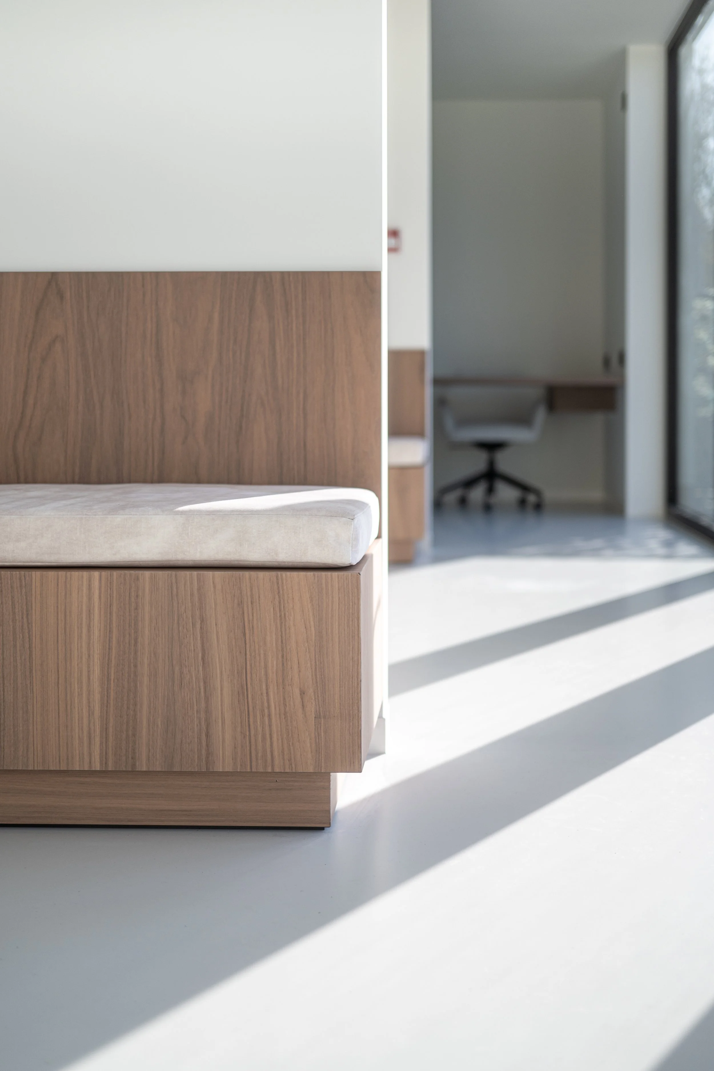 Interieur met een houten bedrand, een matras en een bureau met een bureaustoel in een lichte kamer met veel natuurlijk licht.