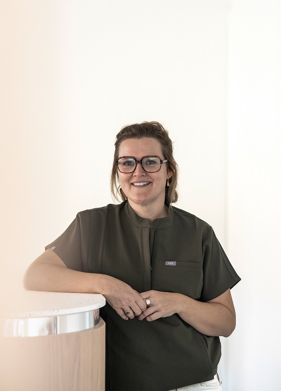 Vrouw met bril en oorbellen, glimlachend, staand naast een witte balie, in een professionele omgeving, draagt een donkere blouse.
