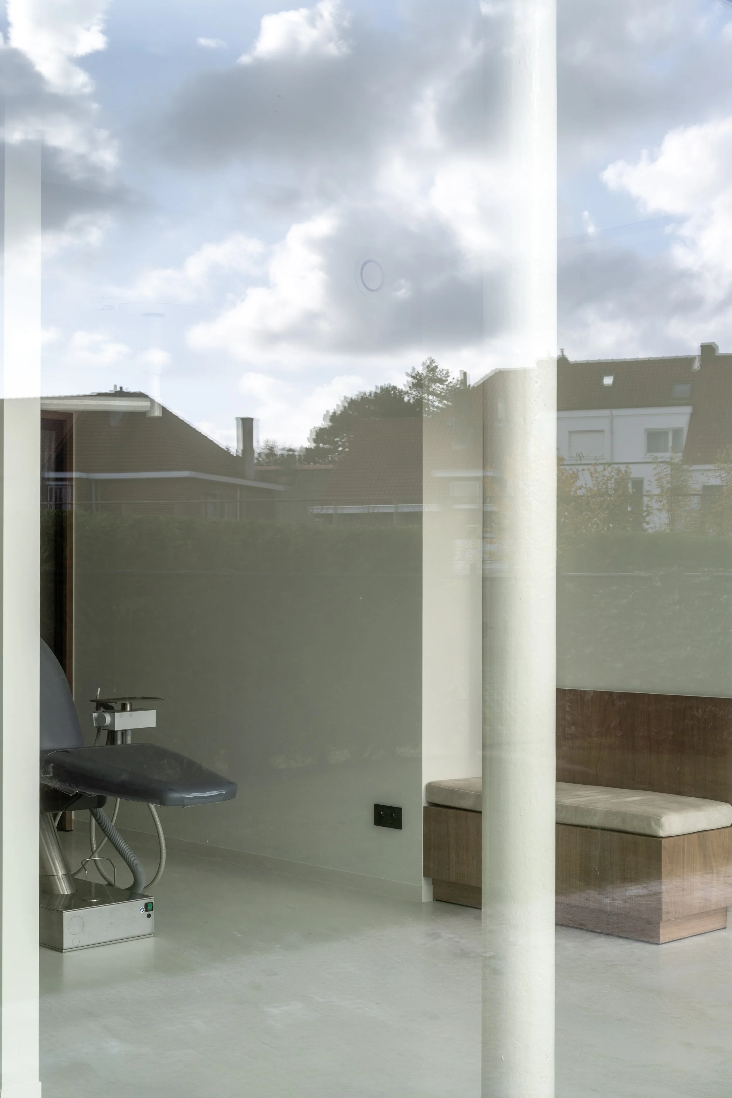 Binnenkant van een ruimte die mogelijk een medische of therapeutische setting is, met een stoel, een apparaat op een poot en een beige bankje, zichtbaar door een grote raampaneel.