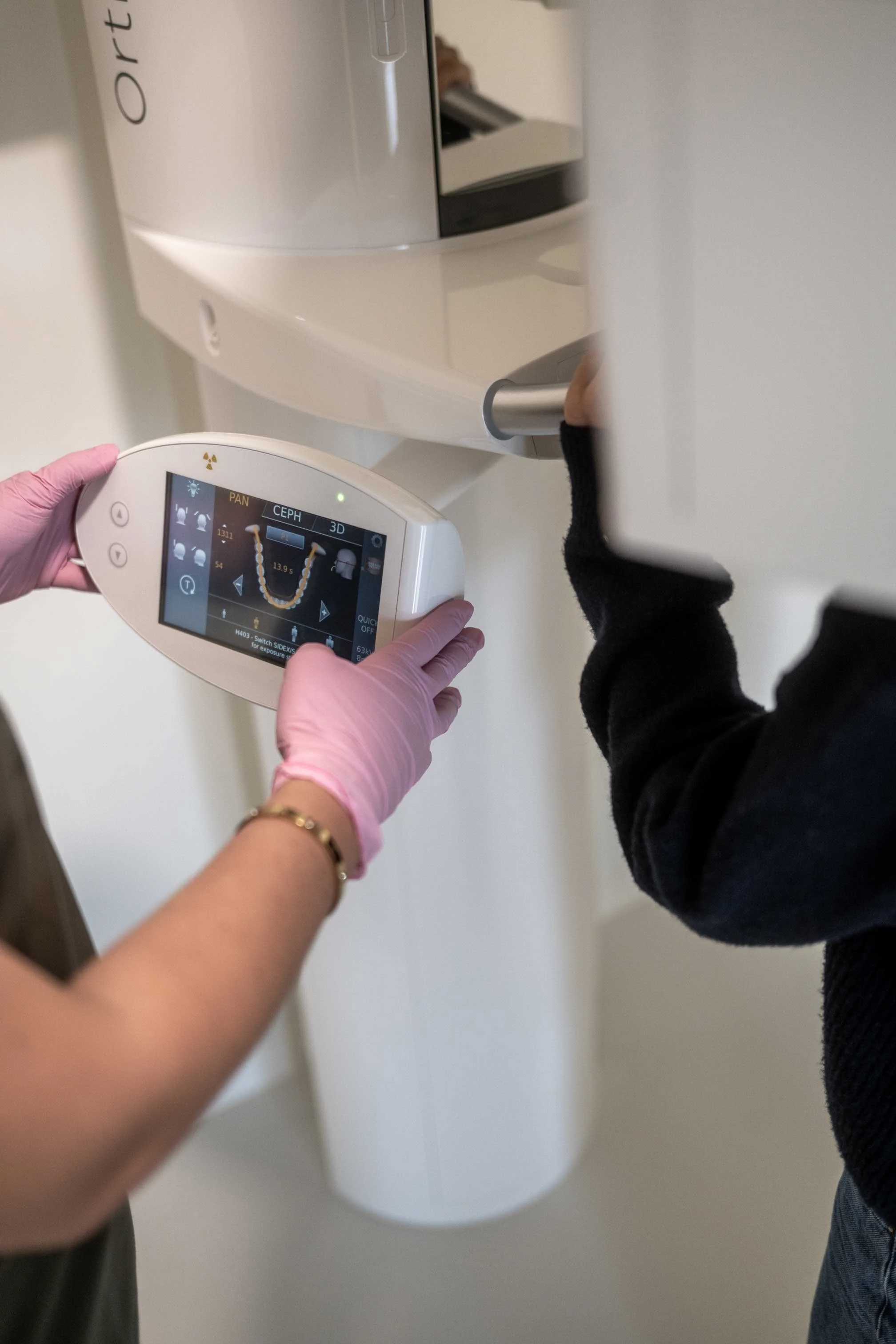 Persoon in roze handschoenen bedient een medisch apparaat dat gebruikt wordt voor medische tests, terwijl een kind met zwarte jas zich vasthoudt aan een grijs handvat.