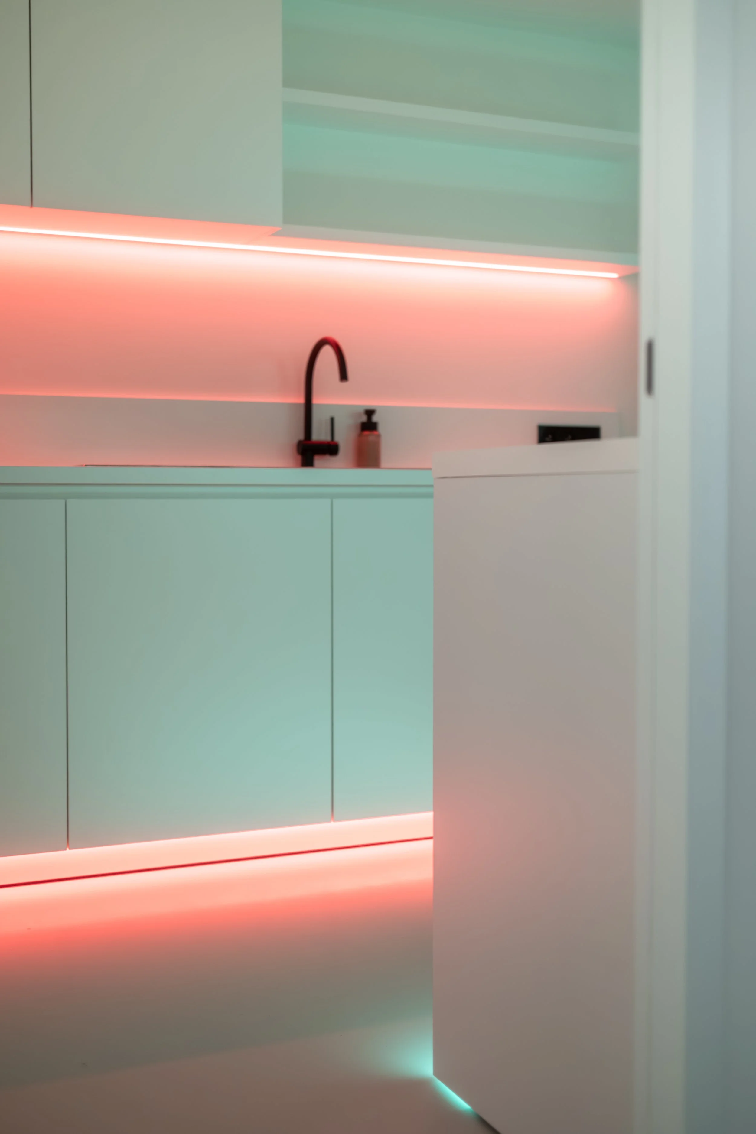 Een hedendaagse minimalistische keuken met vlakke witte kasten en een zwarte kraan. De bovenkasten zijn groen met ingebouwde verlichting die een roze-oranje gloed uitstraalt.
