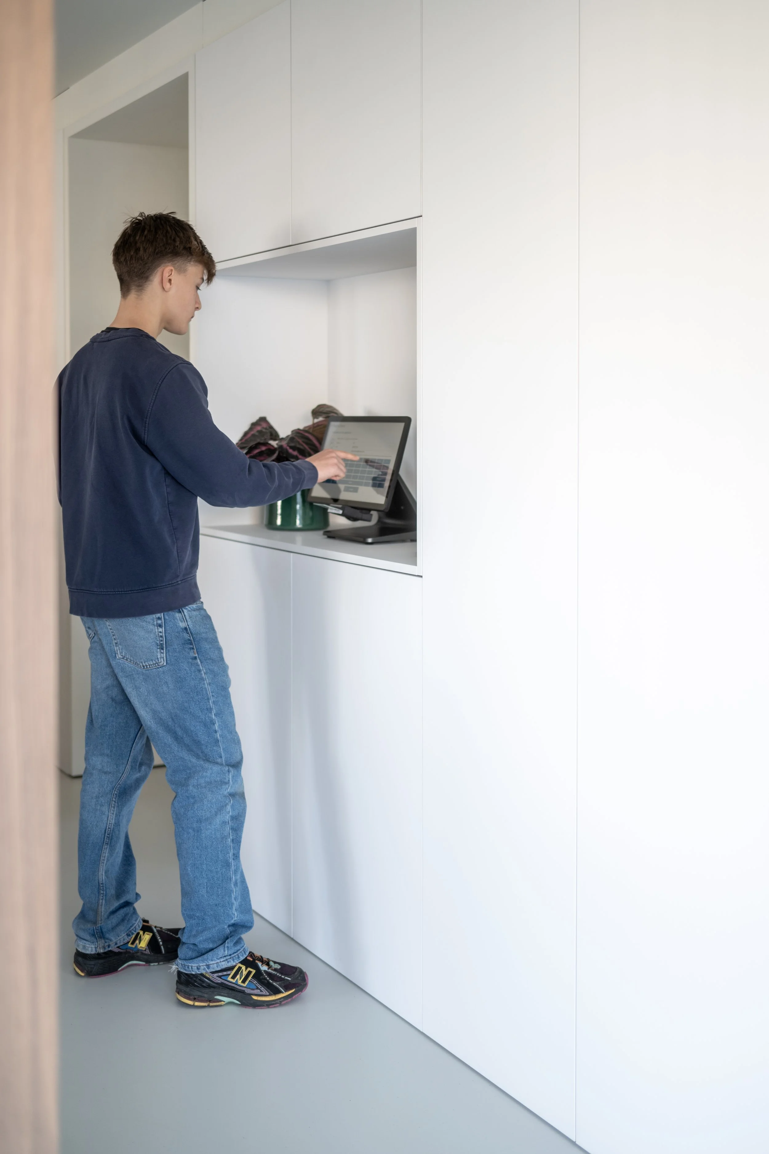 Jongen met kort haar, zittend in een witte kamer, kijkt naar een apparaat op een bureau, draagt een navyblauwe jas en blauwe jeans, en zwart-Nieuw Balance sneakers.