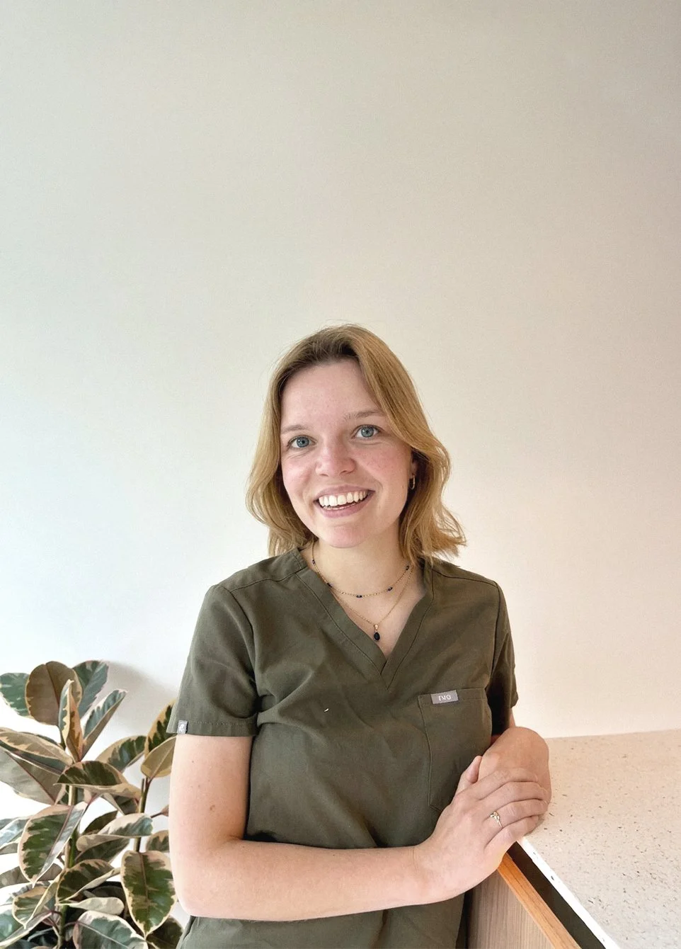 Vrouw met lichtbruin haar in groene sportoutfit die glimlacht en leunt tegen een tafel, met een planten in de achtergrond, in een lichte kamer.