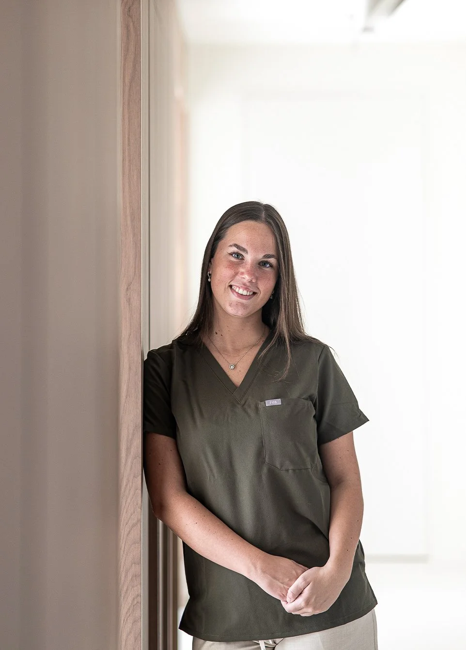 Vrouw in medische outfit die glimlacht en leunt tegen een muur in een lichte, eenvoudige ruimte.