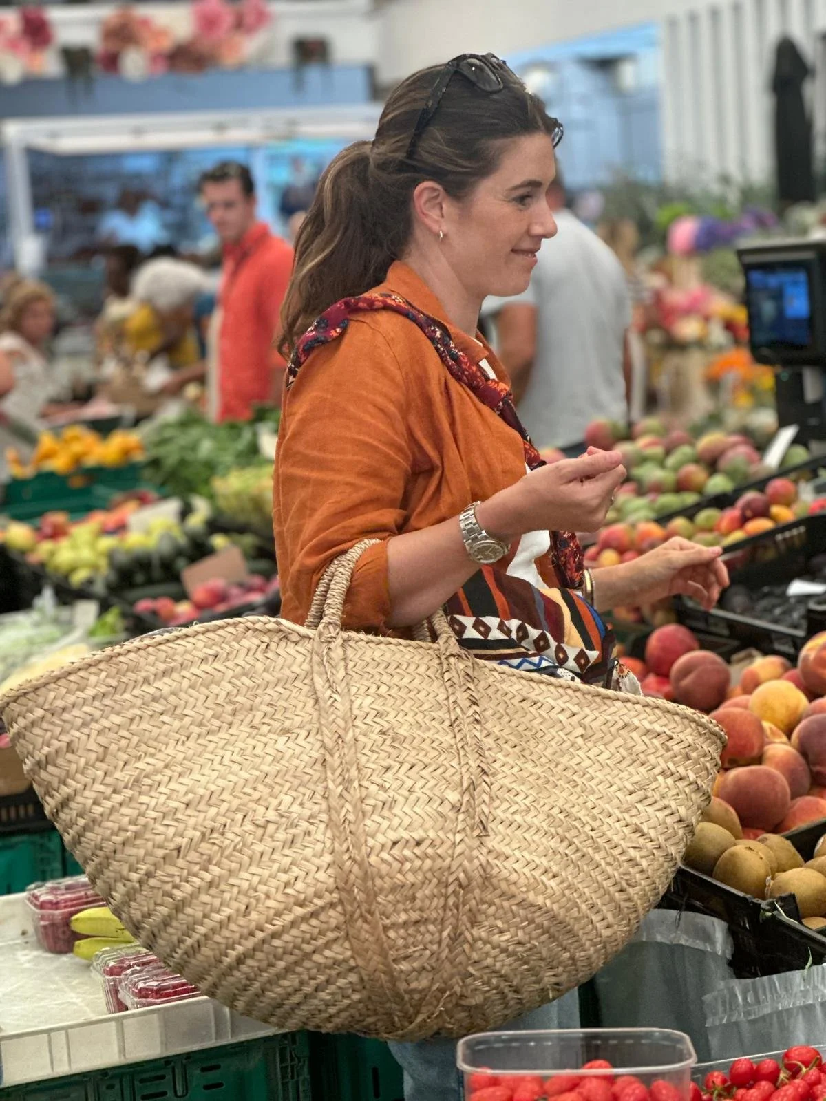 Mujer en un mercado de frutas, con un bolso de paja, mirando la fruta.