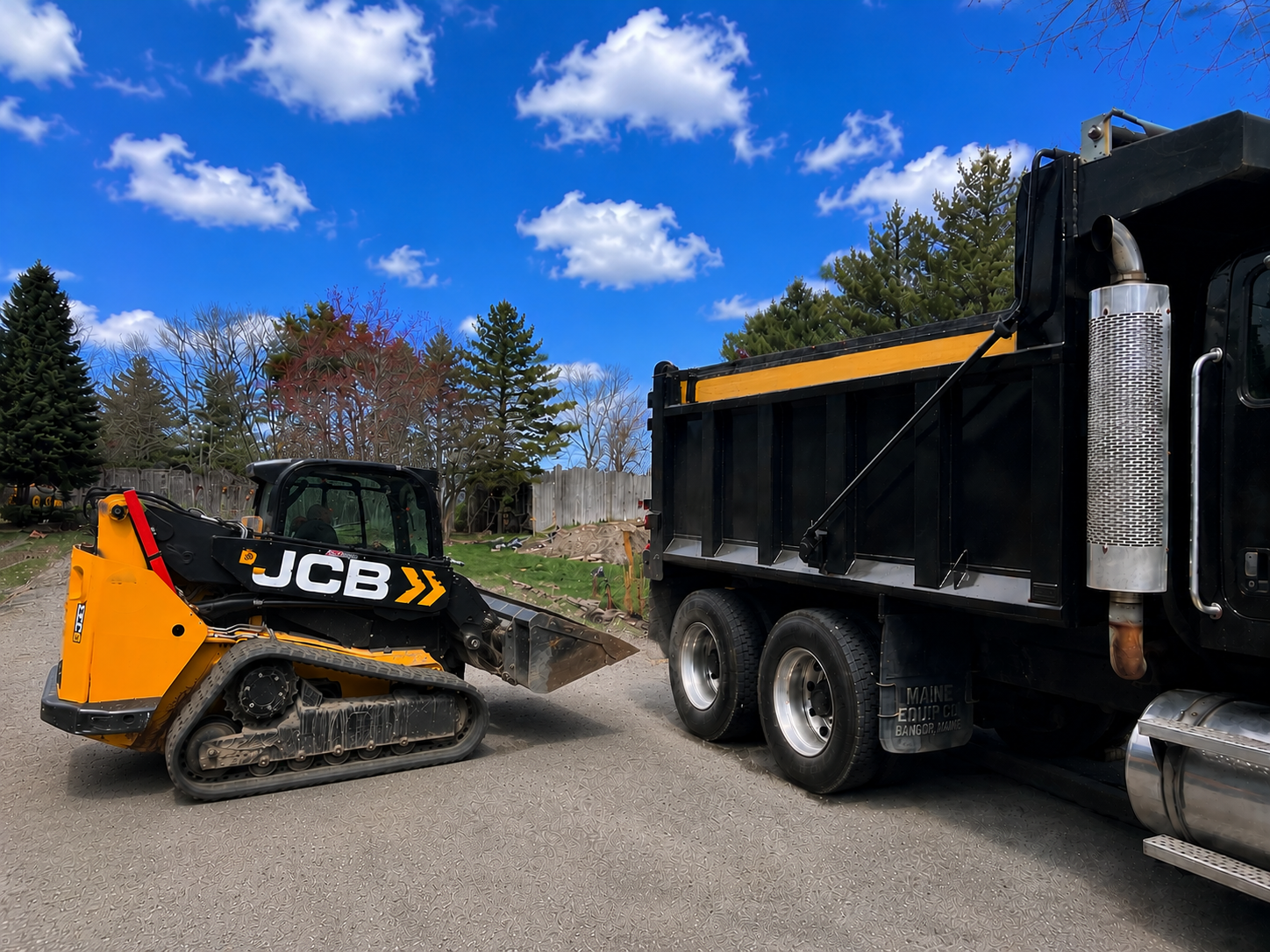 A construction site with a miniature JCB excavator and a large black truck parked on a gravel surface, with trees and a cloudy sky in the background.