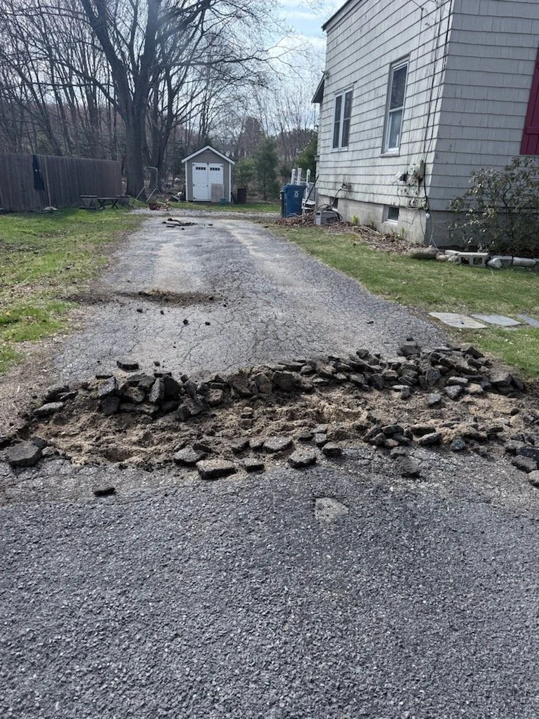 A partially dug and broken asphalt driveway with loose rocks and dirt overlaying it, leading to a white house on the right and a small shed in the background, with bare trees and cloudy sky.
