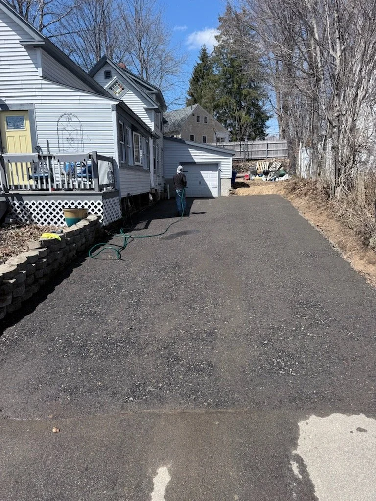A person watering the newly laid gravel driveway in a suburban backyard during daytime, with a two-story house, trees, and a fence in the background.