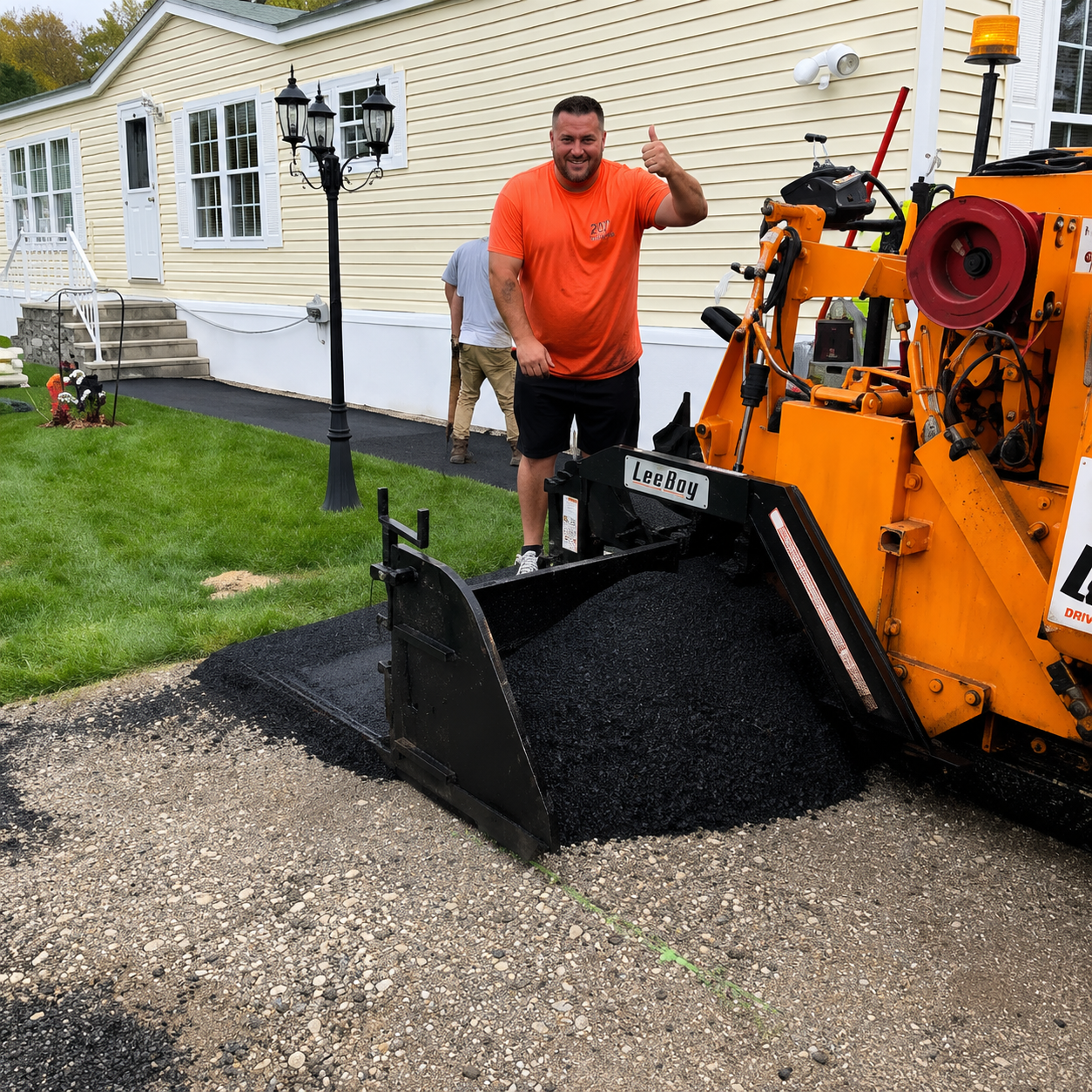 A man in a red T-shirt and black shorts smiling and giving a thumbs-up next to a large orange asphalt paver machine. Behind him, another person is working on the driveway. The setting is a residential yard with a cream-colored house, white steps, a b