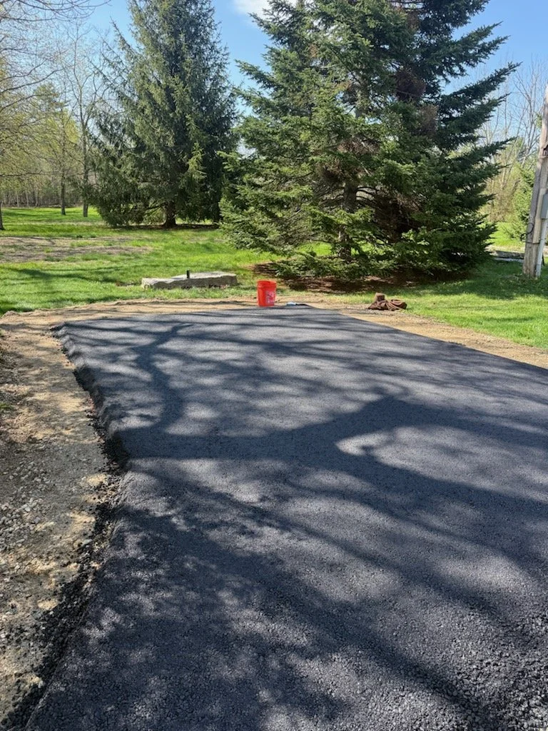 Freshly paved asphalt in a backyard with trees casting shadows, a red bucket, and a pair of brown shoes on green grass.