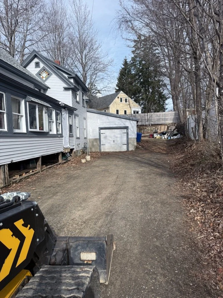A gravel driveway in front of a white house with several windows and a garage, with leafless trees and a neighboring house in the background.