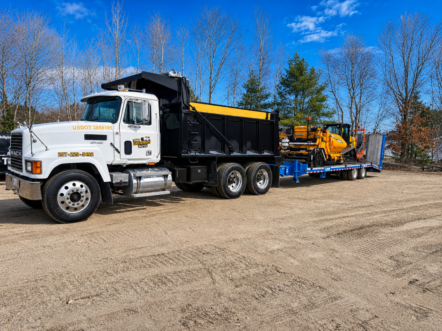 A large flatbed truck parked on a dirt lot, carrying construction equipment including a yellow compactor and a hydraulic excavator, with leafless trees and a partly cloudy sky in the background.