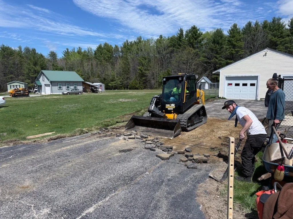 People repairing a damaged driveway with a small bulldozer and hand tools near a white garage in a rural area with houses and trees in the background.