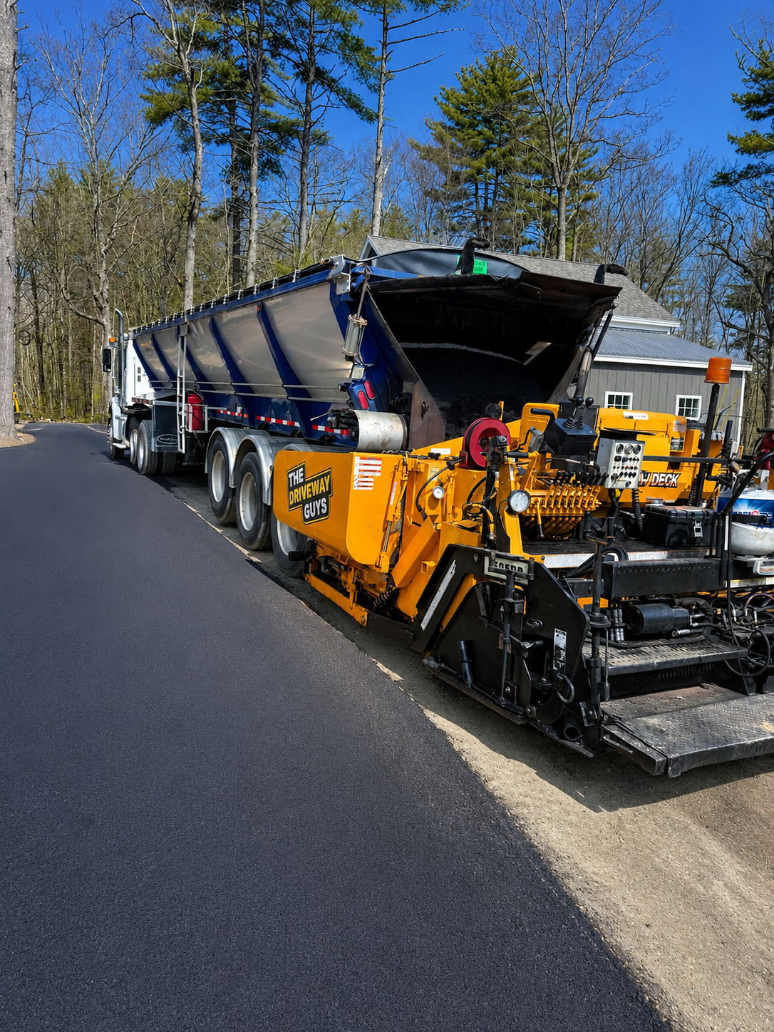 A road paving machine with yellow and black colors, labeled 'The Driveway Guys,' is laying fresh asphalt on a residential street. The machine's hopper is filled with black asphalt, and the surrounding area shows some trees and a gray house.