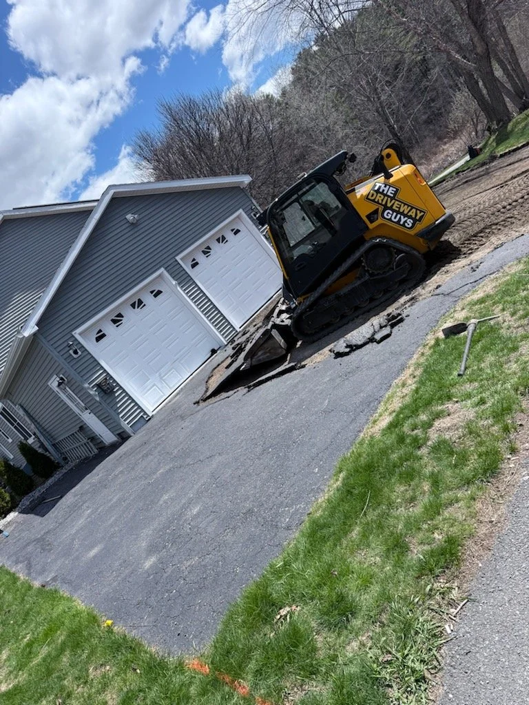 A small excavator with the label 'The Driveway Guys' working on divided asphalt driveway in front of a gray house with white garage doors. The area has some dirt and grass, and the sky is partly cloudy.