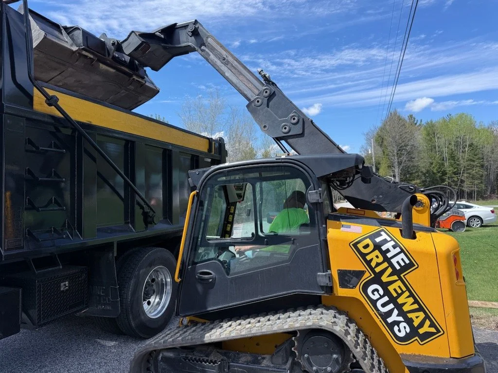 A vehicle with a large black and yellow sign reading 'The Driveway Guys' parked on a grassy area. There is also a compact construction or utility vehicle with tracks and a hydraulic arm, located next to the larger vehicle.