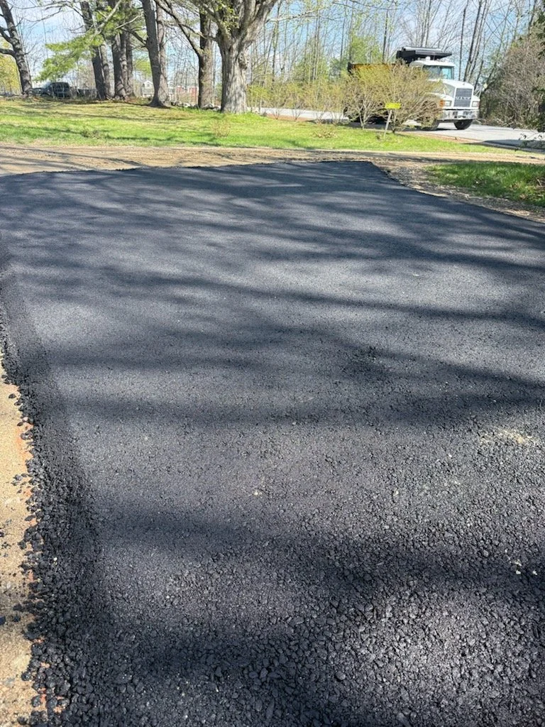 Freshly paved black asphalt driveway with shadows of trees cast across it in a residential yard, with a street and some trees and a white truck in the background.