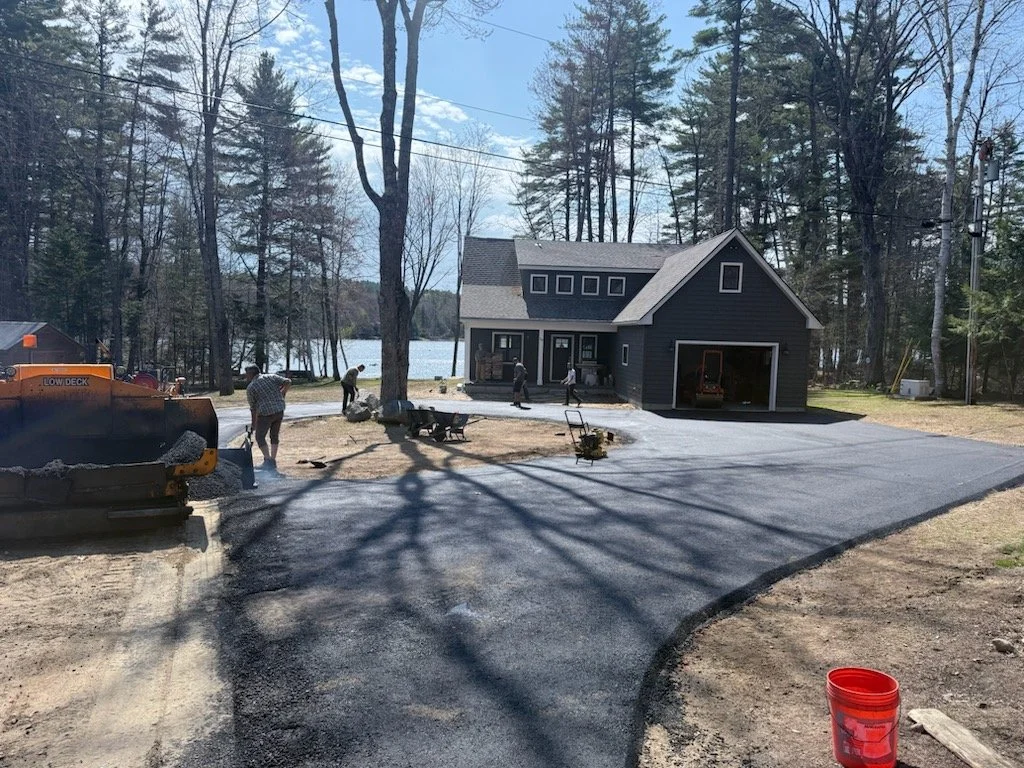 A newly paved driveway in front of a dark gray house with a garage, situated in a wooded area by a lake, with several workers and construction equipment present.