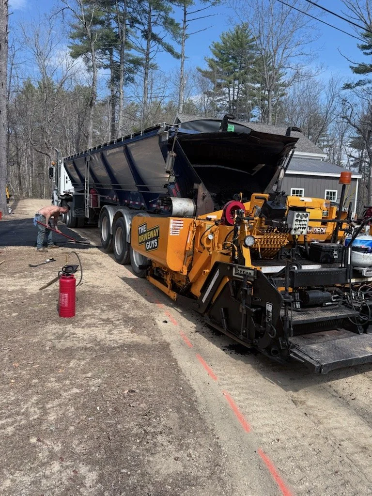A construction worker operating a large asphalt paving machine on a road, with a dump truck nearby and trees in the background.