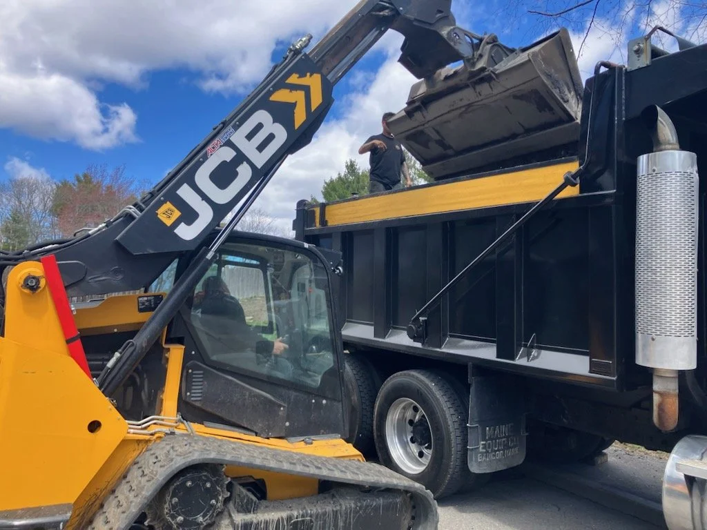 A construction worker operating a JCB backhoe loader and a large dump truck with a man standing inside the truck bed.