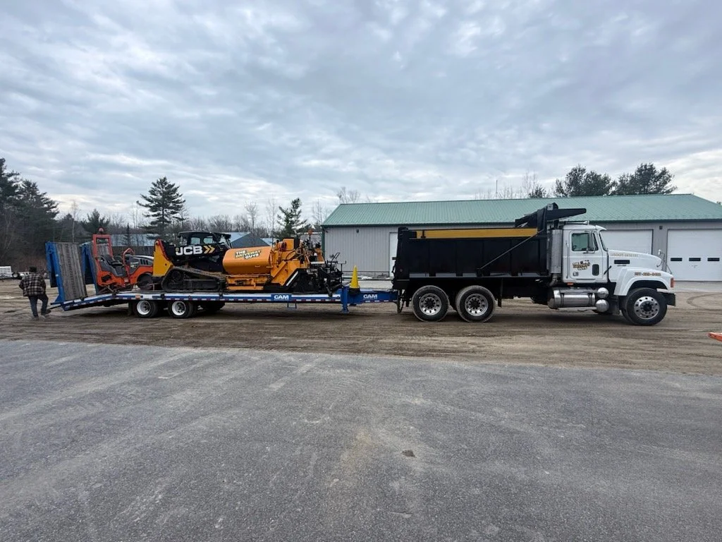 A white truck hauling a flatbed trailer with construction equipment, including a small yellow UCB excavator on the trailer, in a rural area with a building and trees in the background.