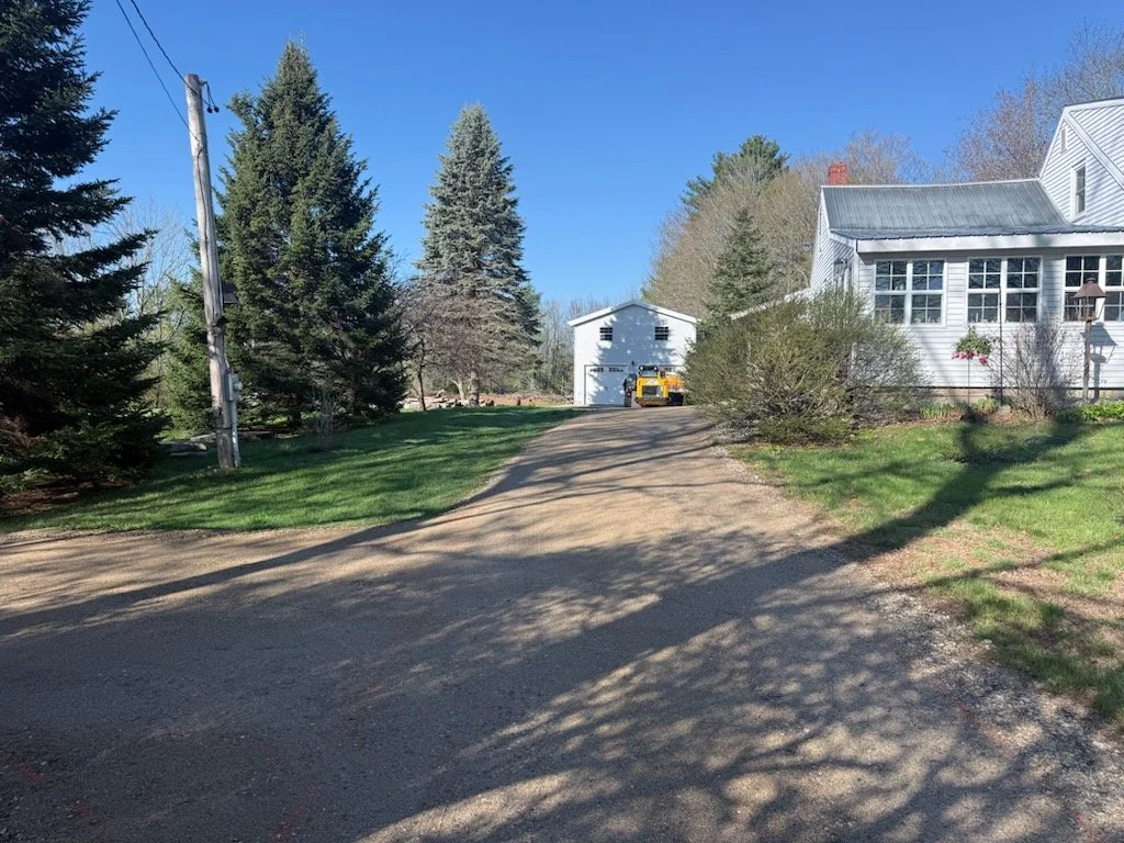 A dirt driveway leading to a white garage with a yellow vehicle parked in front, surrounded by trees and suburban houses on a sunny day.