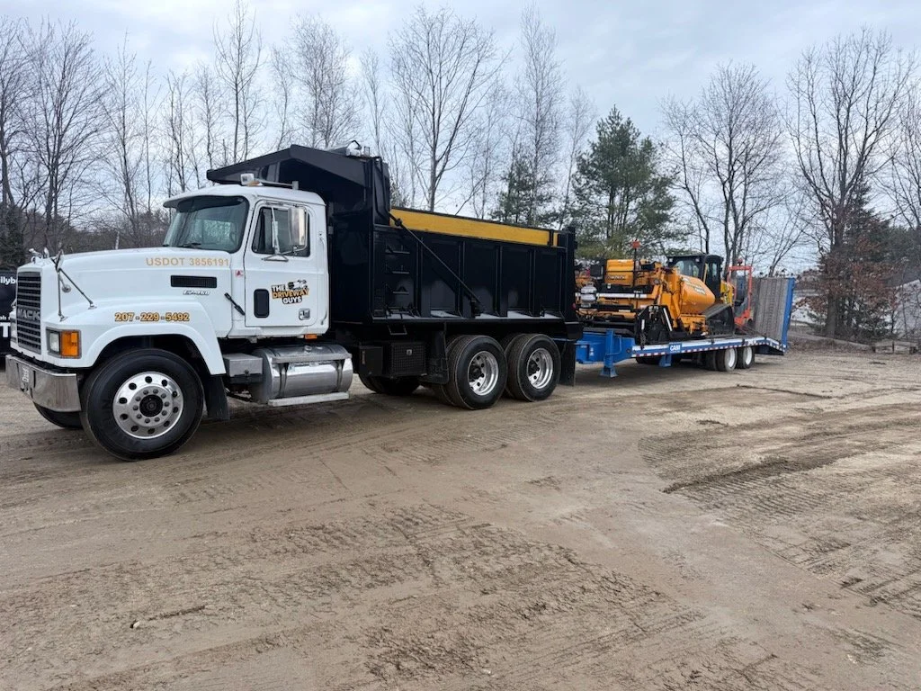 A flatbed truck parked outdoors on a dirt surface, carrying construction equipment including bulldozers and excavators, with leafless trees in the background.