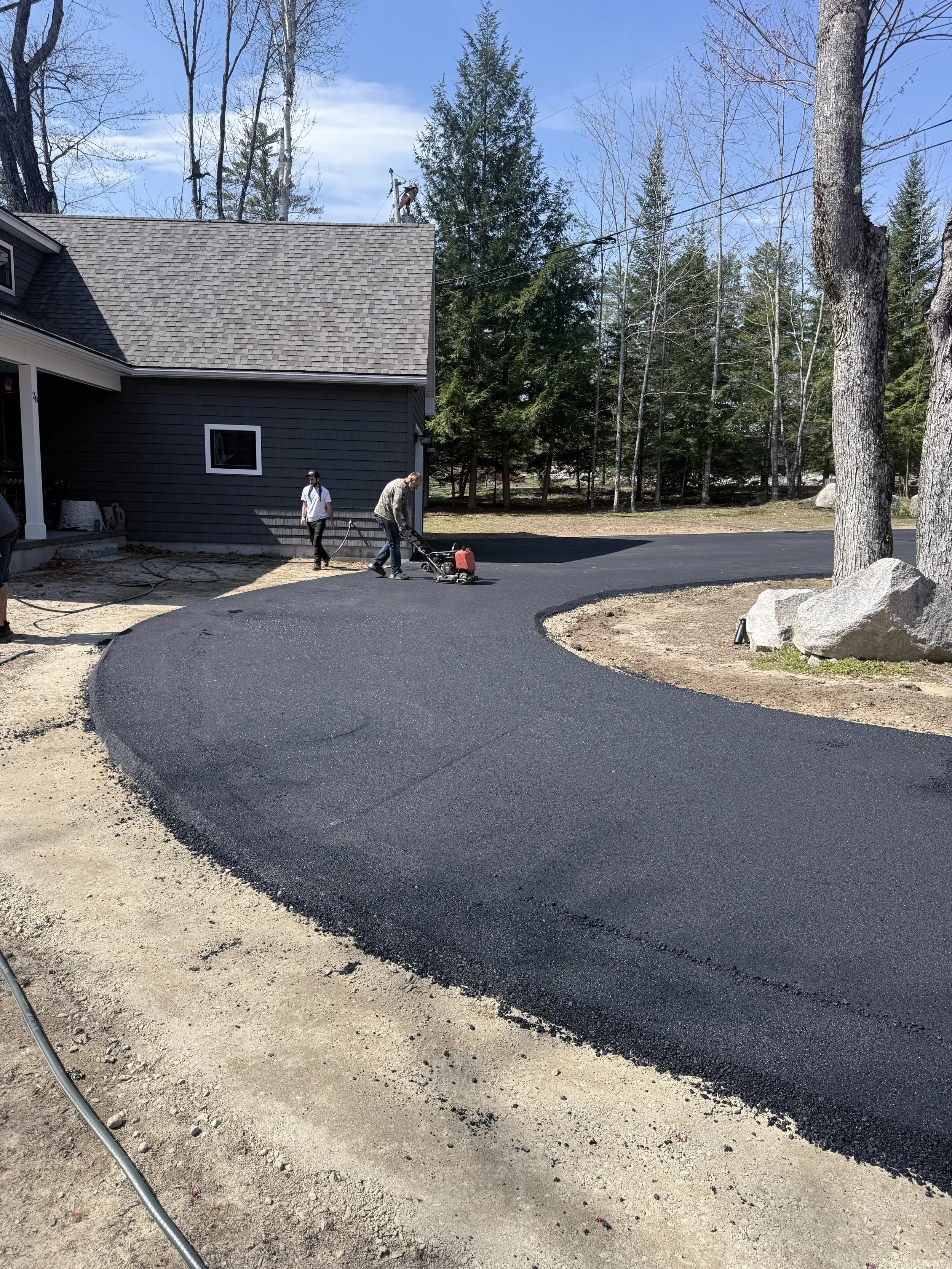 Two men are working on paving a curved driveway with fresh black asphalt in a residential area. One man is using a tamper, and the other is working nearby.