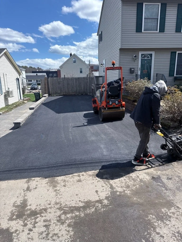 A person in a dark jacket and hoodie using a machine to smooth fresh asphalt on a driveway outside a house, with a roller compactor parked nearby.