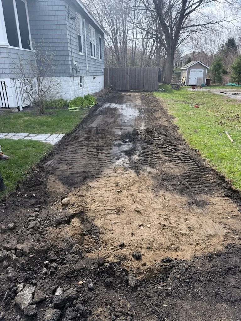 A backyard with a dirt pathway being prepared for construction, bordered by grass and a house on the left, with a wooden fence and trees in the background.