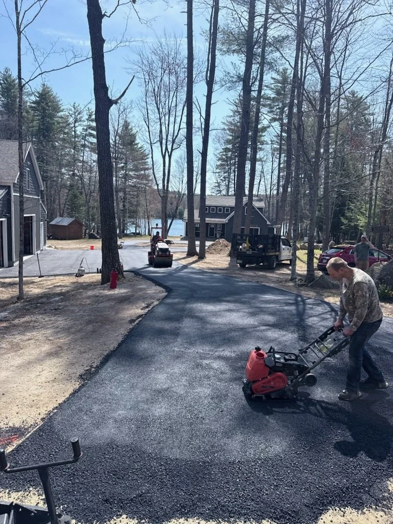 Man operating a plate compactor on a newly paved asphalt driveway in a residential neighborhood with trees and houses, during daytime.