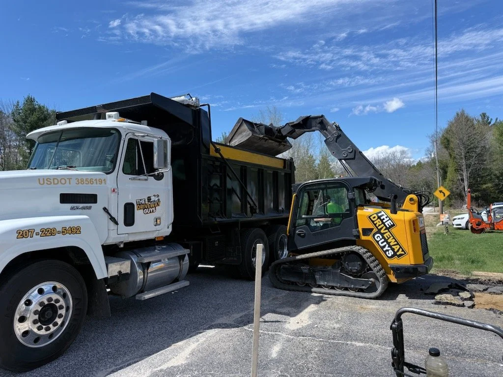 Construction work involving a large white truck and a small yellow and black excavator on a paved road with some dirt and broken asphalt, under a partly cloudy blue sky with trees in the background.