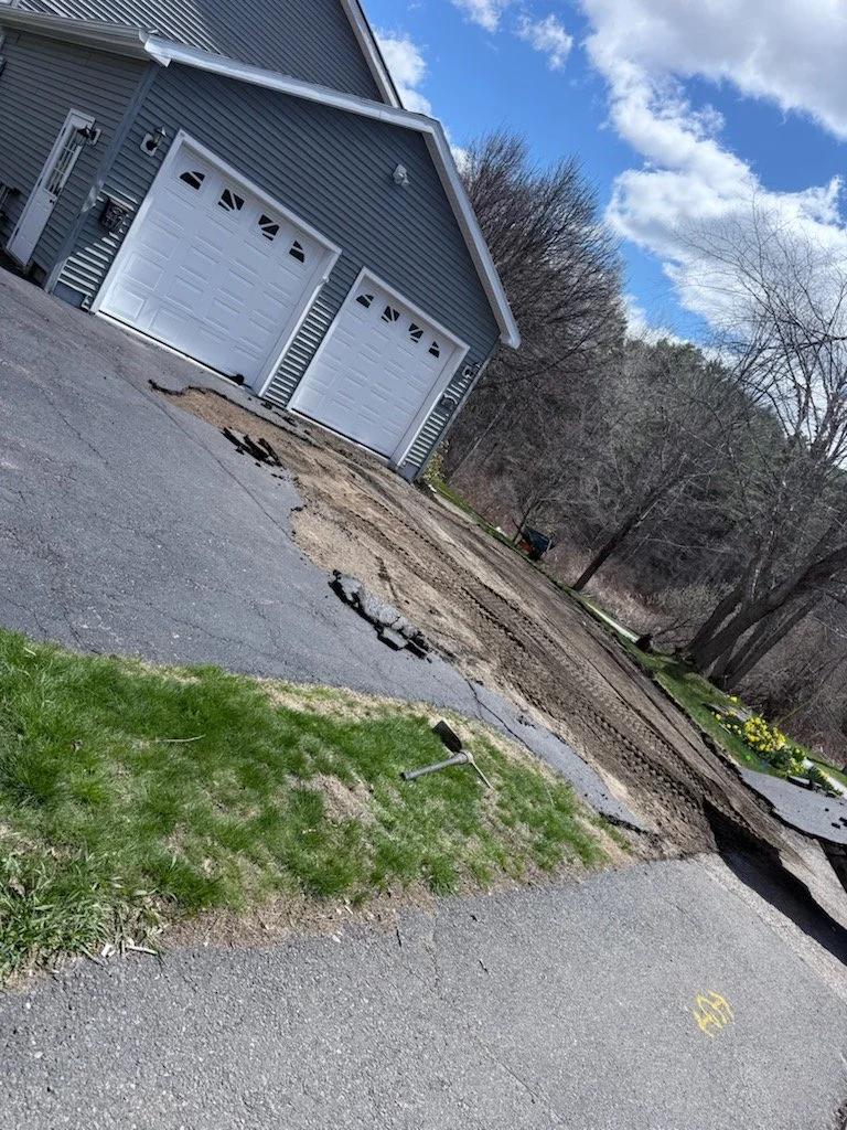 Driveway being repaired with tire marks, a hammer on the grass, and a house with closed garage doors in the background.