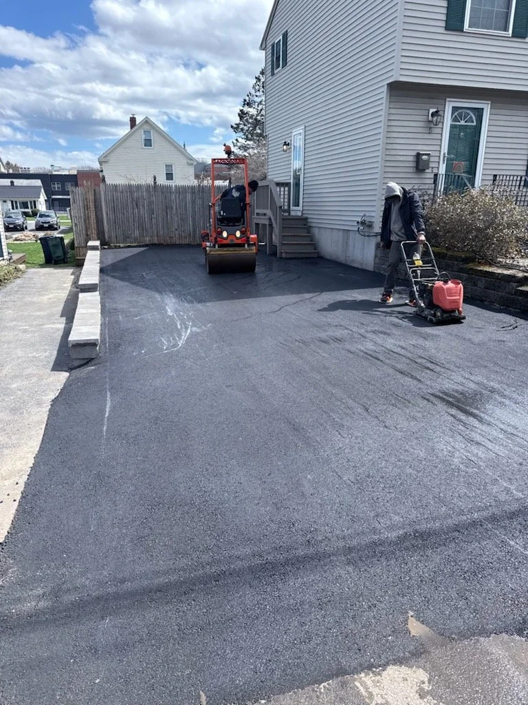 A residential driveway being freshly paved with black asphalt. A worker is with a power compactor, and a roller machine is nearby. A house with siding is on the right, with steps leading to a door. The sky is partly cloudy.