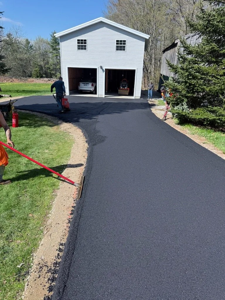 Workers paving a new asphalt driveway leading to a white garage with two open doors, parked cars inside, and green trees in the background.