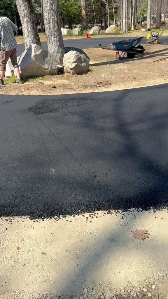 A freshly paved black asphalt road with no visible markings, neighboring a patch of bare light-colored dirt, with trees and rocks in the background, and a wheelbarrow near the road.
