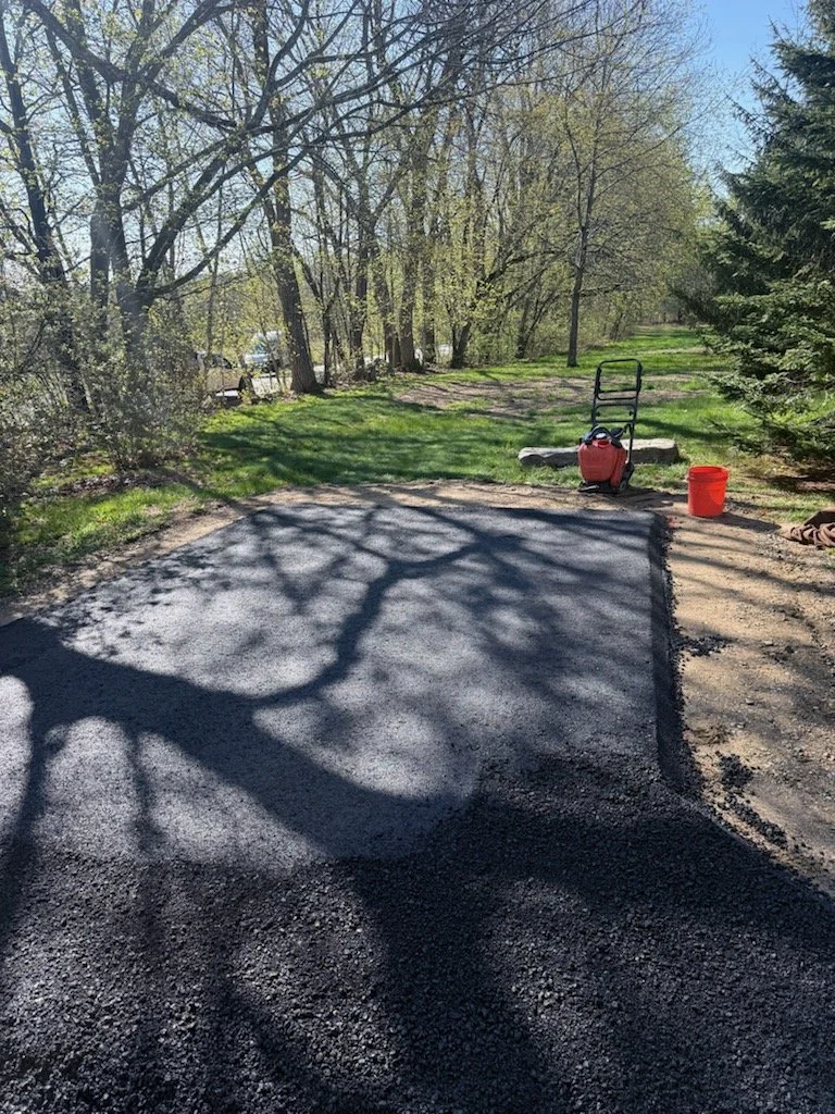 A backyard scene with a patch of fresh asphalt being laid, surrounded by grass and trees. Garden tools, including a wheelbarrow, bucket, and a lawn roller, are visible nearby.