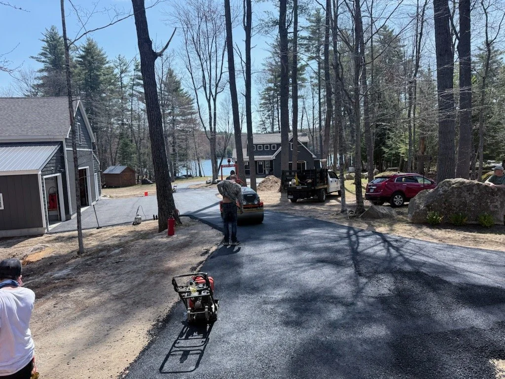 People are paving a driveway near a lake in a wooded area, with construction equipment and parked cars around.