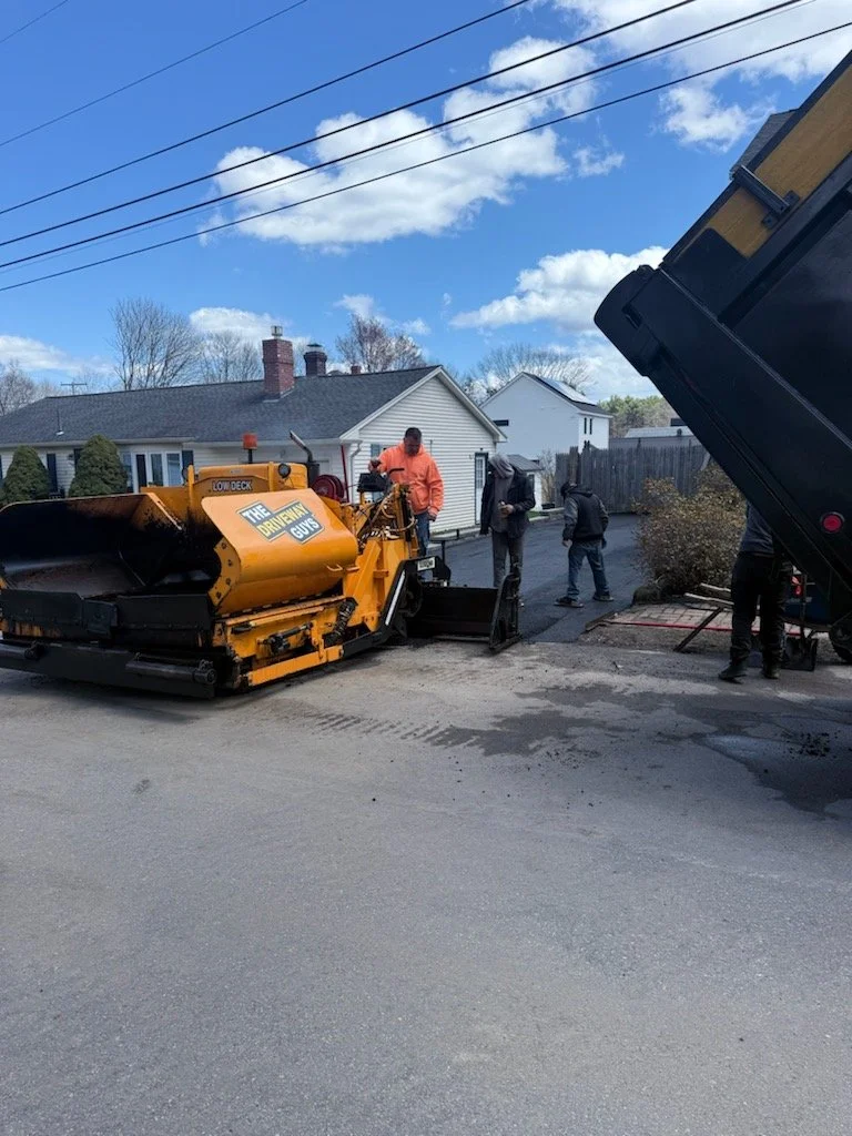Workers operate asphalt paving equipment on a residential street, with some workers standing nearby and a house with a white exterior in the background.