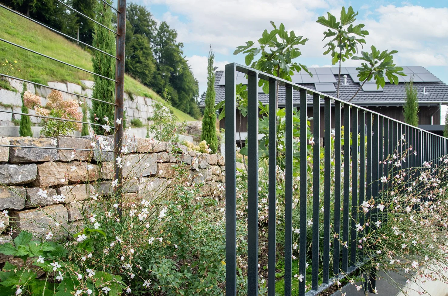 Garten mit Steinmauer, schwarzen Metallzaun, Blumen und Bäumen, in der Nähe von einem modernen Haus mit Solarpanelen auf dem Dach.